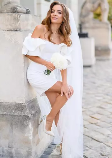 front-view-charming-blonde-bride-with-curly-hairs-long-veil-wearing-short-wedding-dress-with-puffy-sleeves-holding-bouquet-while-leaning-stone-wall-raising-leg-heels-outdoors_8353-12479