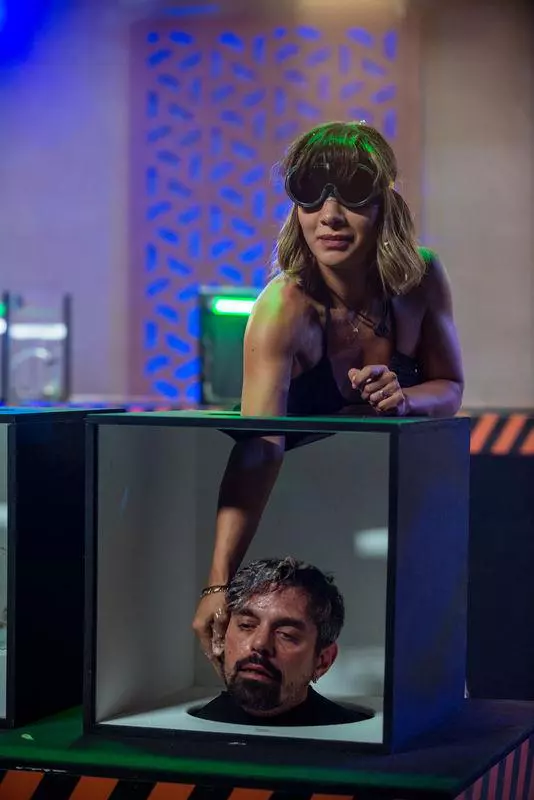 Anca Munteanu, cu ochelari negri, asemănători unor ochelari de protecție, își întinde mâna într-o cutie neagră deschisă în partea de sus. Pare concentrată în timp ce atinge ceva necunoscut. În fața ei, într-o altă cutie identică, se vede capul lui DOC. Lumina ambientală are nuanțe de albastru și verde, sugerând o atmosferă de competiție sau spectacol de televiziune.