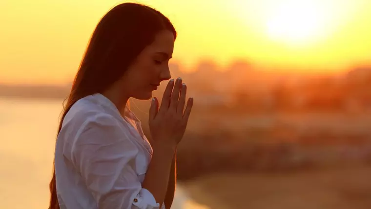 femeie care se roaga cu ochii inchisi si mainile impreunate, la apus de soare