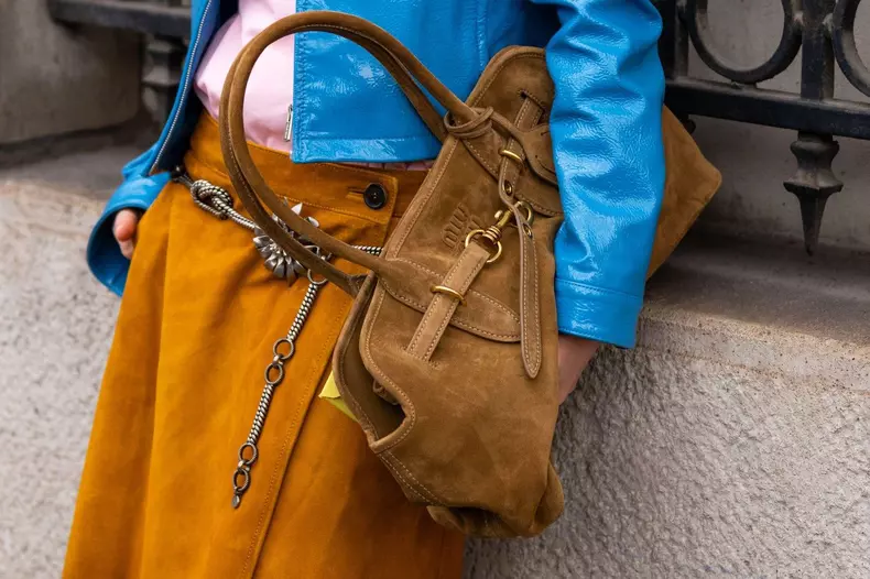 Paris, France - March 11, 2025: Street style. Brown suede Miu Miu handbag with gold hardware, worn with blue patent jacket and ochre skirt