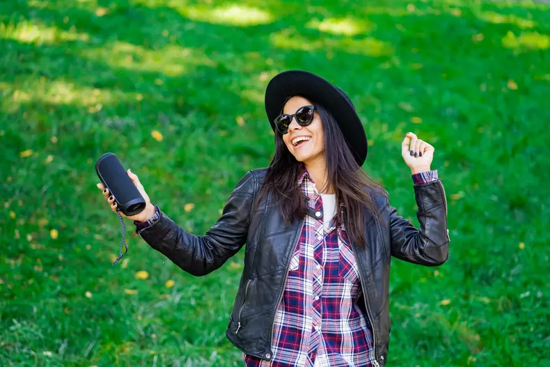 Happy mixed race young woman listening to music with wireless portable speaker. Hispanic hipster girl dancing to rhythm and singing along melody in the park