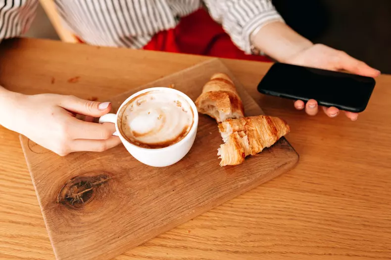 Crop photo of cup with coffee and french croissants ion the table on wood desk. Young lady with smartphone having breakfast on cafe