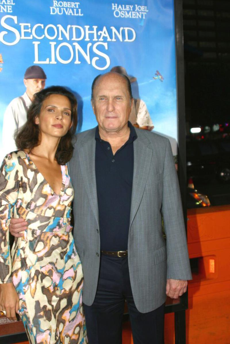 Robert Duvall și soția sa Luciana Pedraza la premiera filmului Secondhand Lions din 2003. Robert Duvall poartă un costum gri cu cămașă albastră, iar Luciana poartă o rochie neagră cu decolteu în V. Cei doi zâmbesc fotografilor pe covorul roșu.