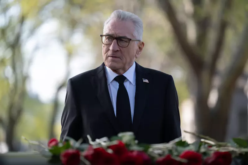 . Robert de Niro, în costum și cravată, stând în fața unei coroane, din serialul Netflix Zero Day.