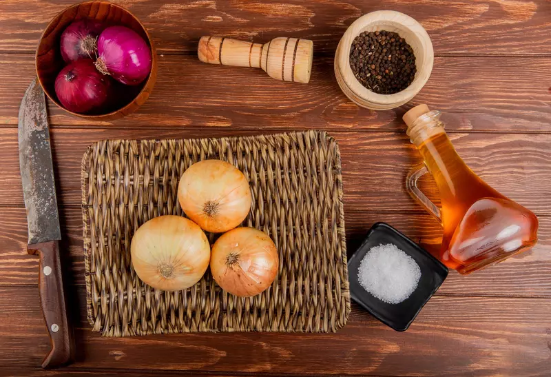 top view of onions as red and sweet ones in bowl and in basket plate with butter salt black pepper seeds and knife around on wooden background
